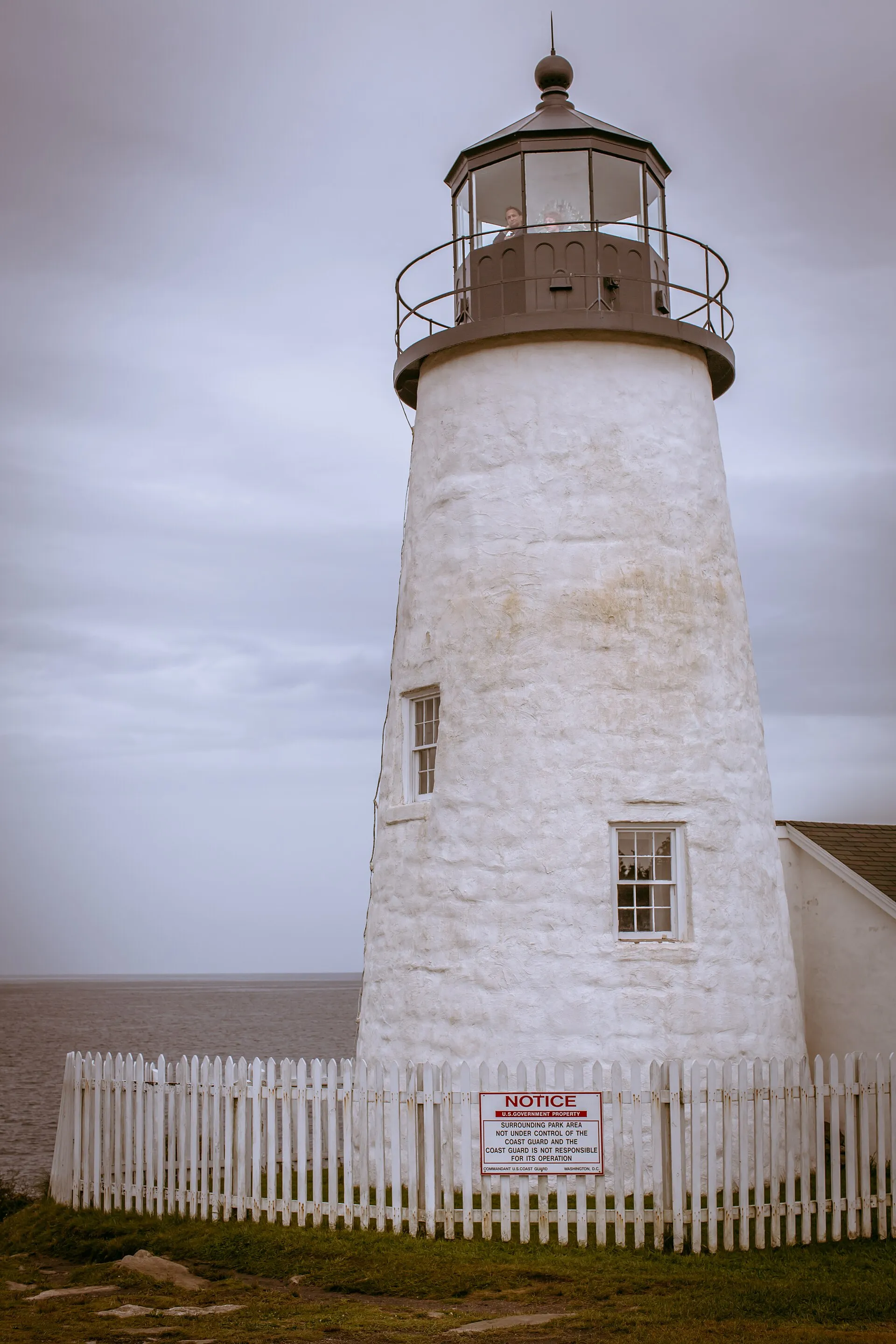 Pemaquid Point Light, a whitewashed tower behind a white picket fence, with visitors standing in the lantern room at the top